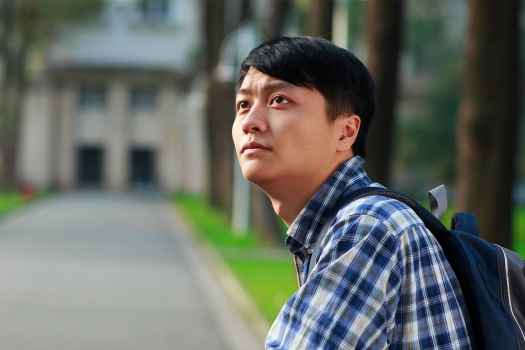 Asian man with backpack and plaid shirt looking up.