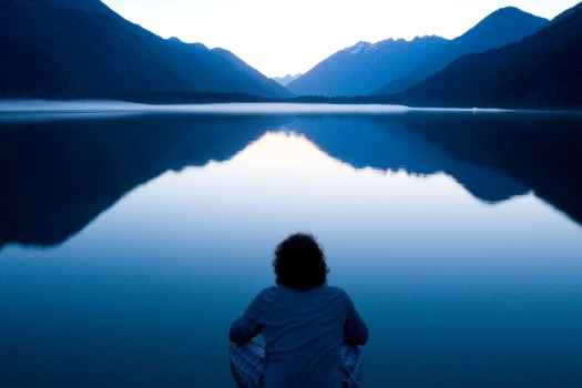Man sitting looking at a still lake and mountains in the distance