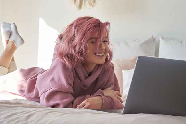 Girl with pink hair watches laptop, lying on bed