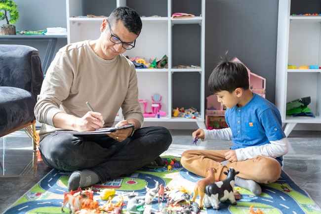 Child plays on rug with therapist sitting taking notes