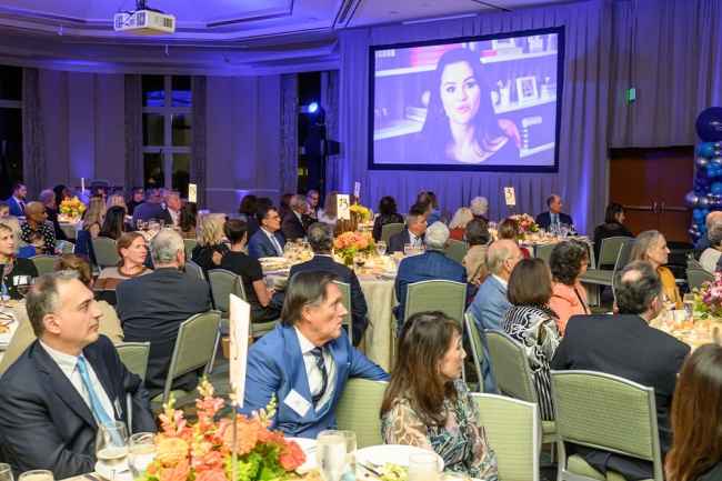 People sitting at tables looking at a screen with a woman talking