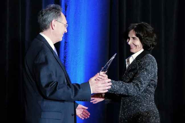 One person hands another person a clear glass award in front of dark blue curtain