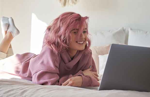 Girl with pink hair watches laptop, lying on bed