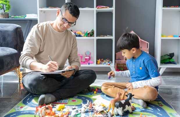 Child plays on rug with therapist sitting taking notes