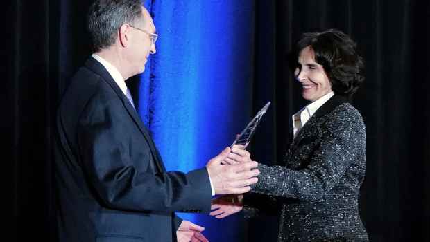 One person hands another person a clear glass award in front of dark blue curtain